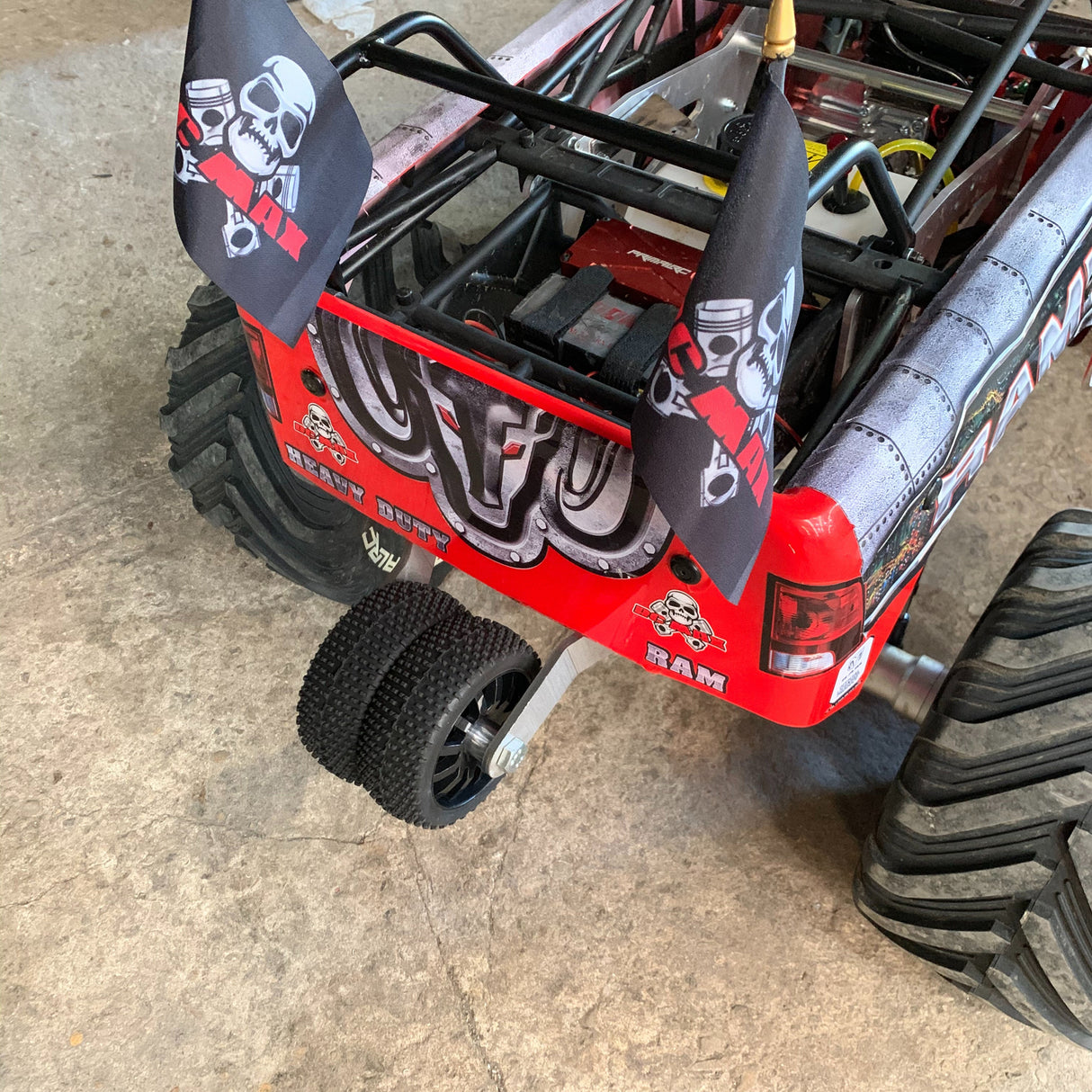 A remote control toy accessory, a wheelie bar, is shown mounted on the back of a red and black remote control truck with a billet centre shaft and RC wheels.