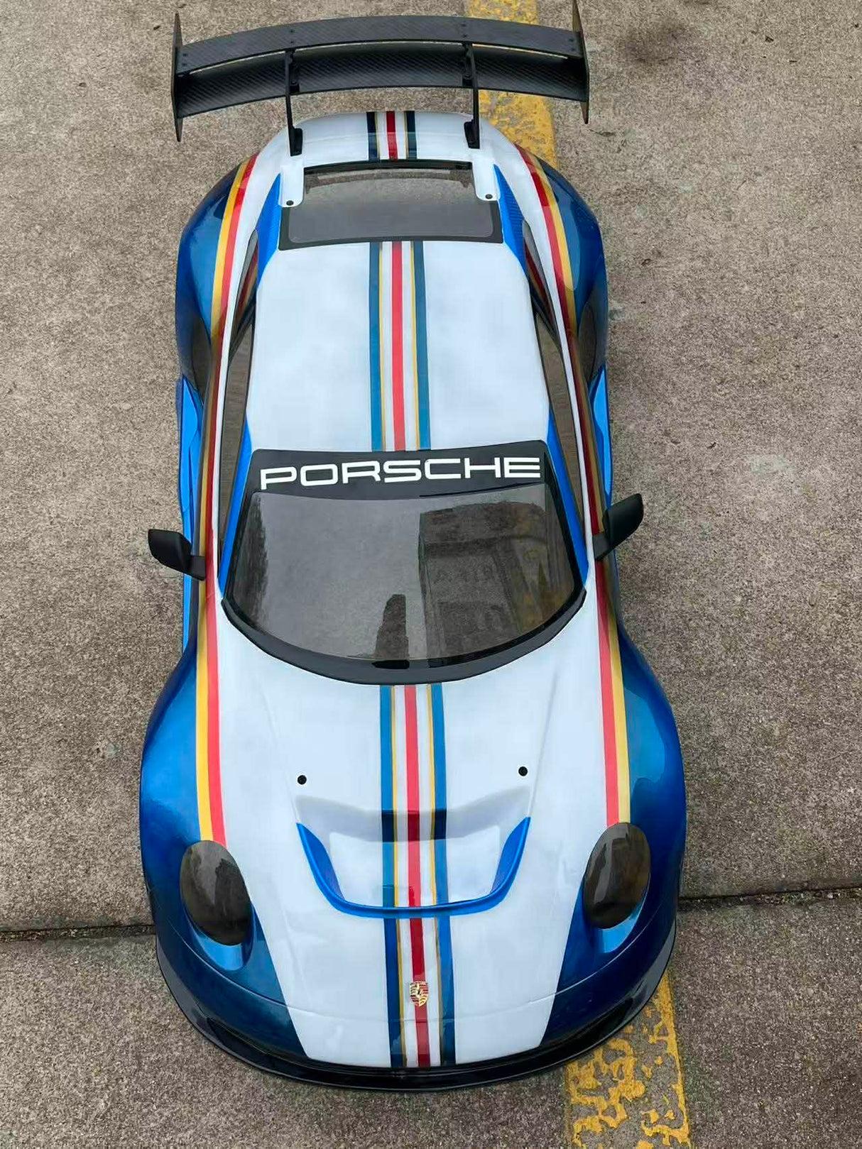 Top view of a Porsche car with a white hood featuring red, blue, and yellow stripes on a concrete surface.