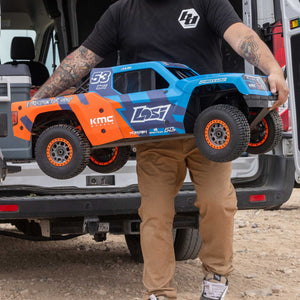 Person holding a blue and orange remote control truck in front of a vehicle