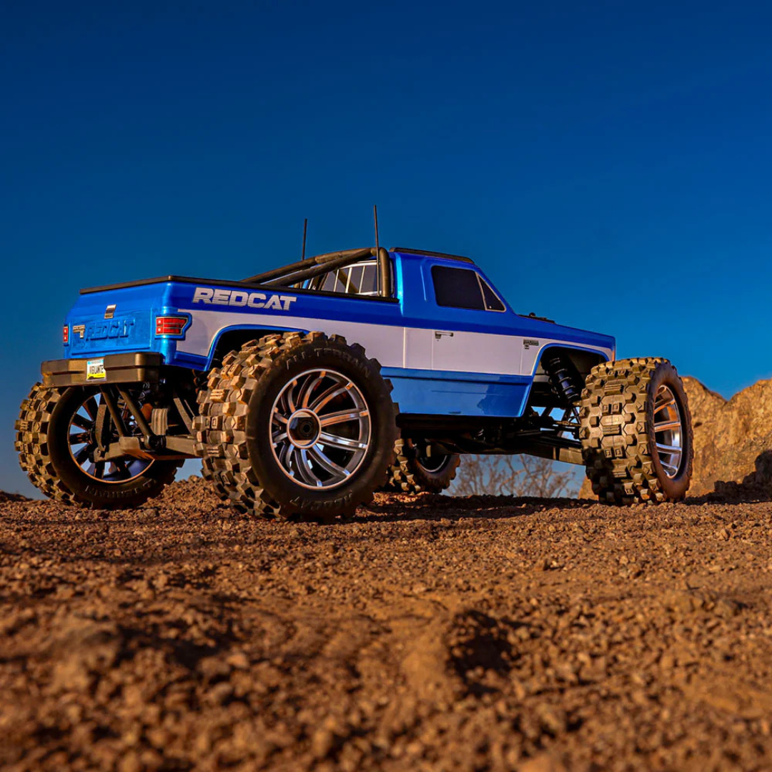 Blue remote-controlled monster truck on a dirt surface with a clear blue sky.