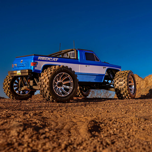 Blue remote-controlled monster truck on a dirt surface with a clear blue sky.